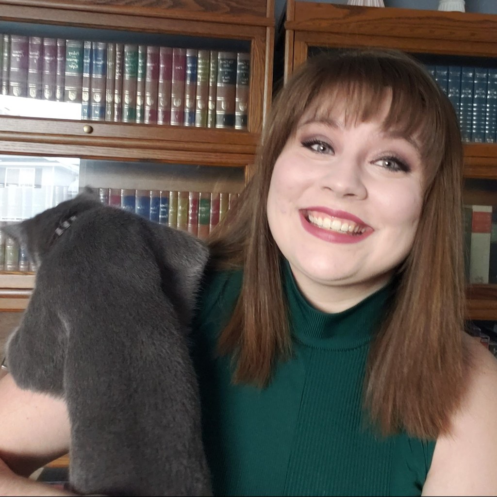 Michelle Smiling at the camera in front of a book shelf with a cat on her shoulder. 